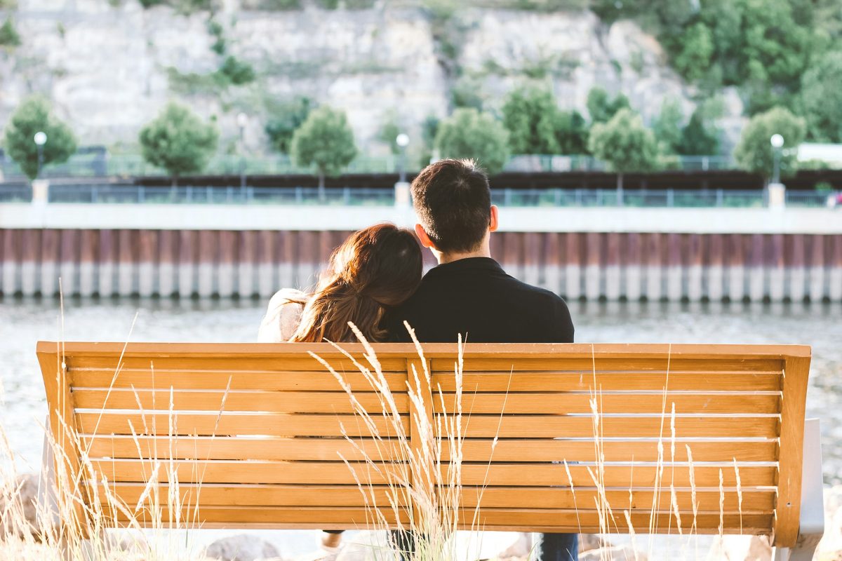 Couple seen from behind on a wooden bench by a river, her head resting on his shoulder