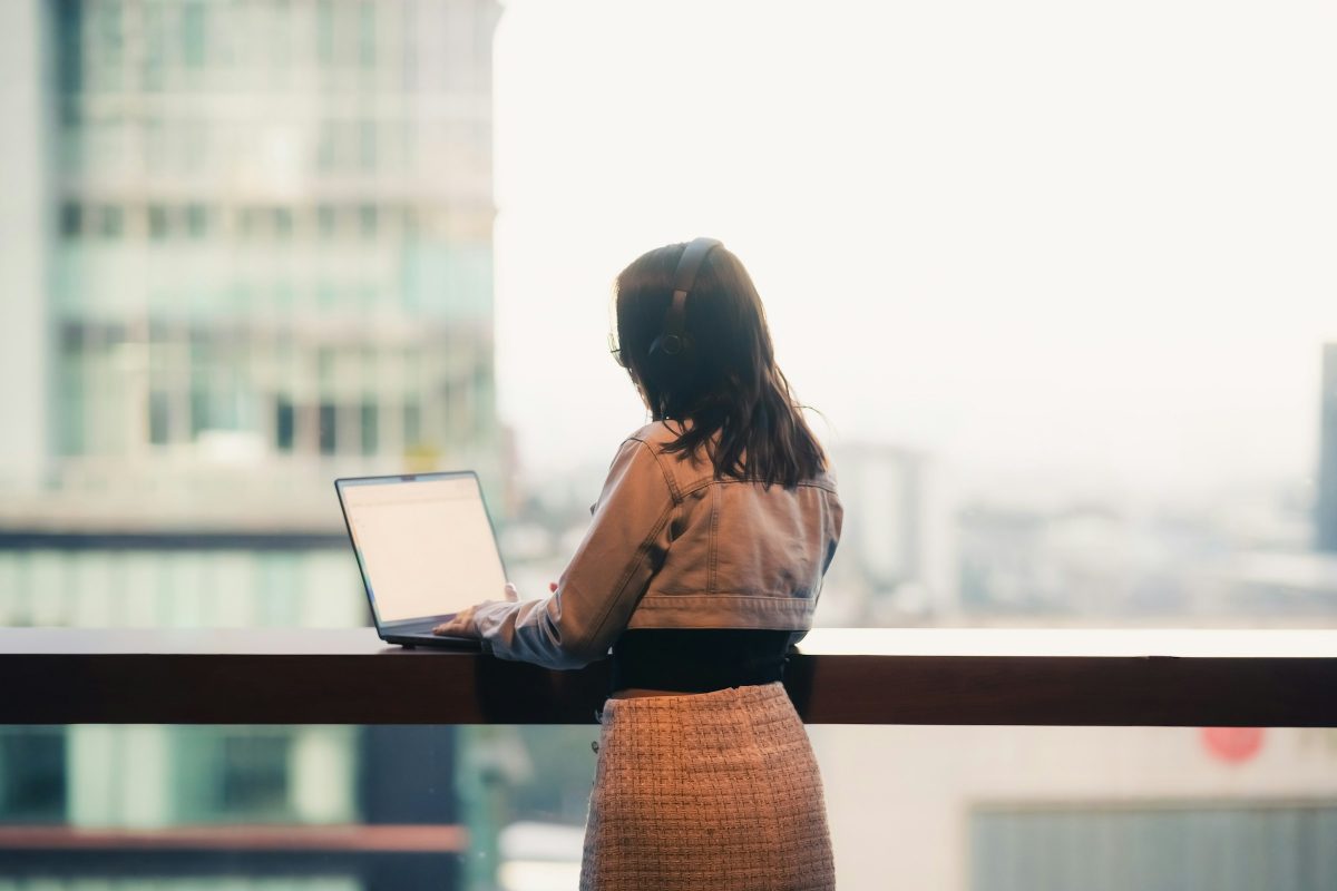 Woman with headphones working on a laptop at a window counter overlooking a city