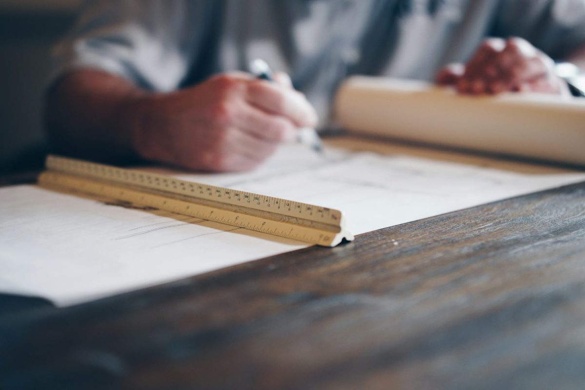 Hands drafting a technical drawing with a pen and scale ruler on a wooden desk