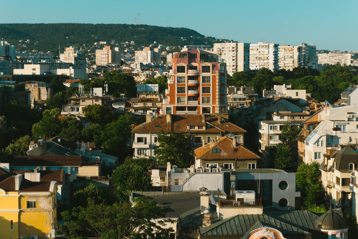 Apartment blocks and tiled-roof houses among green trees in Varna, with a wooded hill behind