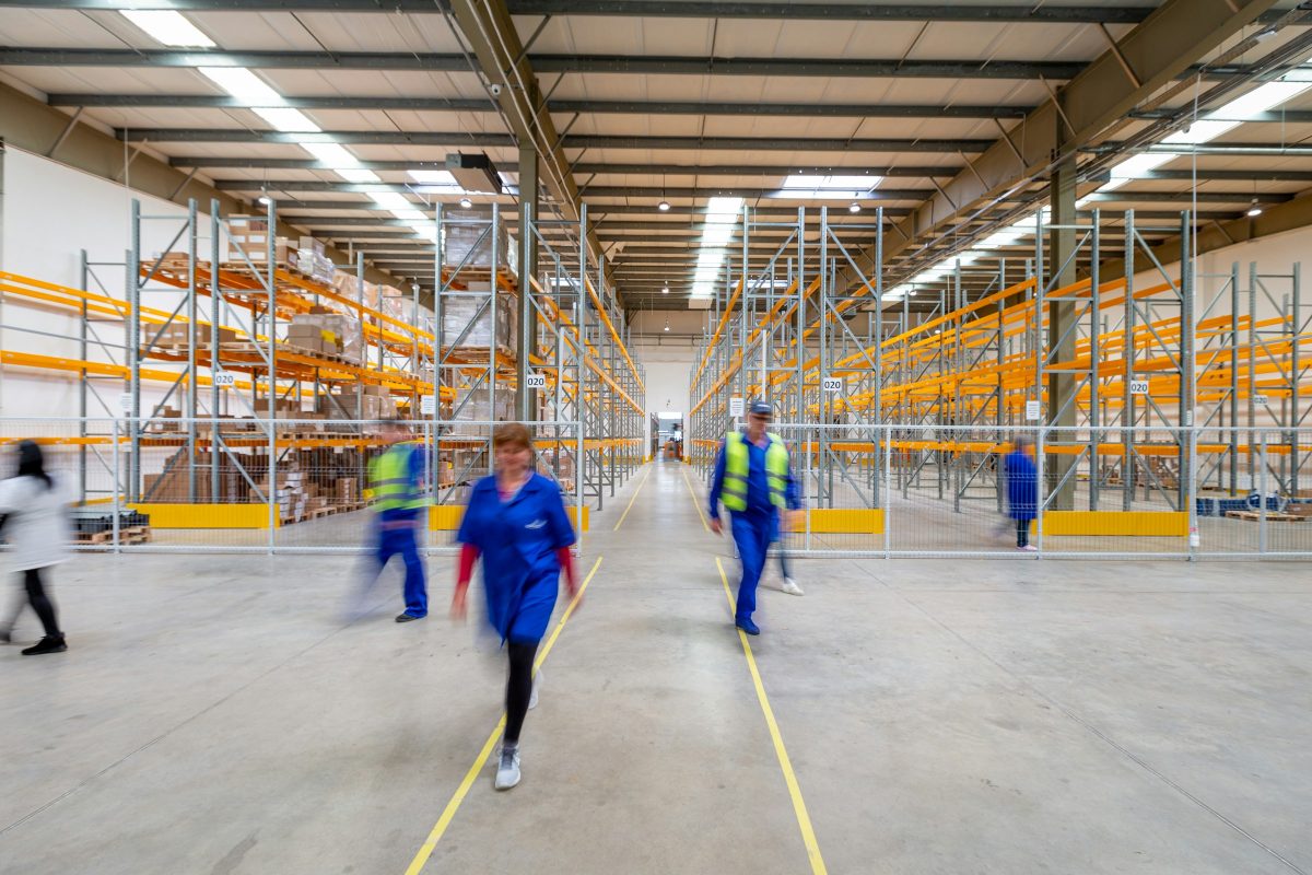 Workers walking along a wide warehouse aisle between tall orange pallet racks